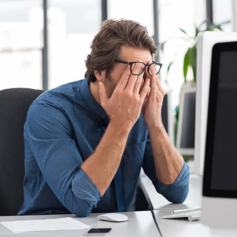 Man with blude long sleeve button up shirt sitting at desk and rubbing his eyes under his glasses in discomfort.